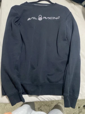 Svart sweatshirt från Sail Racing - Mörkblå sweatshirt från Sail Racing med logga i vitt på ryggen. Klassisk rund halsringning och långa ärmar med ribbade muddar. Perfekt för en avslappnad och sportig stil.