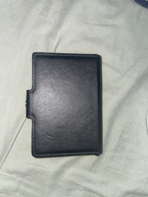 Svart minimalistisk plånbok i läder - Snygg och stilren svart plånbok i slätt läder med minimalistisk design. Plånboken har en kompakt och fyrkantig form, perfekt för kort och sedlar. Passar dig som gillar enkel och modern stil.