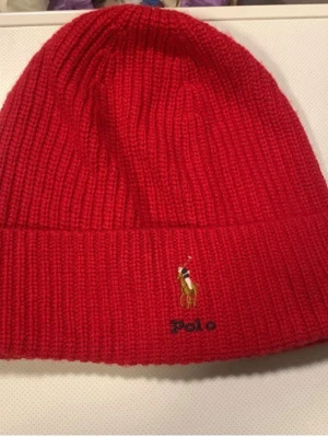 Röd ribbstickad mössa från Polo Ralph Lauren - Säljer en snygg röd ribbstickad mössa från Polo Ralph Lauren med klassisk broderad logga framtill. Mössan har uppvikt kant och är tillverkad i mjukt, värmande material som passar perfekt för kyliga dagar.
