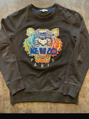 Svart sweatshirt med tiger från Kenzo - Cool svart sweatshirt från Kenzo med ikonisk broderad tiger i flera färger på bröstet och texten 'KENZO PARIS'. Tröjan har rund hals, långa ärmar och ribbade muddar. Perfekt för dig som vill sticka ut med en statement-piece.