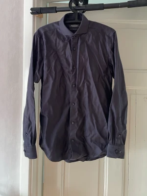 Mörkgrå skjorta från Dressmann - Stilren mörkgrå skjorta från Dressmann, med klassisk krage och knappar framtill. Skjortan har lång ärm med manschett och är tillverkad i mjuk bomull. Endast använd ett fåtal gånger. Lite skrynklig på bilderna men det fixar man med ett strykjärn🤩