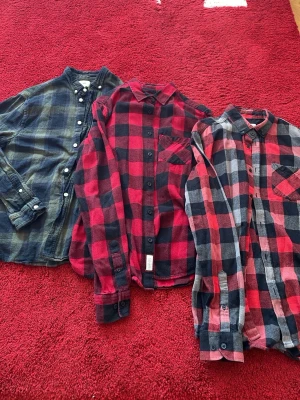 3 rutiga flanellskjortor i olika färger - Tre långärmade skjortor i mjuk flanell, alla med klassiskt rutigt mönster. Två skjortor är röd- och svart-rutiga, en är svart och grönrutig. Alla har knappar framtill och bröstficka. Storlek L i alla. De är från H&M och Lager 157.