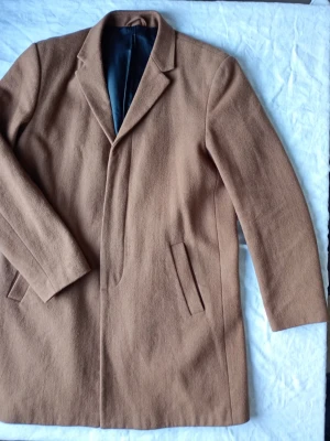 Klassisk kamelfärgad ullkappa - Stilren kamelfärgad kappa i ullmix med enkel knäppning och två snedställda fickor framtill. Kappan har en klassisk krage och svart innerfoder, perfekt för en trendig och tidlös look. Passar till många olika outfits och ger en elegant siluett.