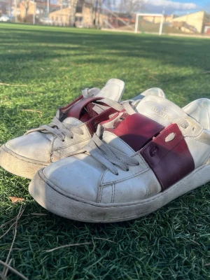 Valentino vita sneakers med vinröda detaljer - Snygga vita sneakers från Valentino med breda vinröda läderdetaljer över mitten och klassiska vita snören. Skorna har en låg profil och är tillverkade i läder med diskret logga på plösen. Perfekta för dig som vill ha en clean men ändå lyxig look.