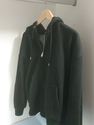 Svart zip hoodie  - En klassisk svart hoodie med dragkedja framtill och stora fickor. Tröjan har huva med dragsko och ribbade muddar vid ärmslut och nederkant. Perfekt för en chill och avslappnad stil. Materialet ser ut mjukt och bekvämt, troligen bomullsblandning.