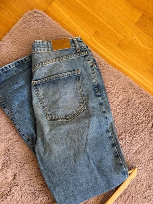 Blå raka jeans från Gina Tricot - Säljer ett par klassiska blå jeans från Gina Tricot, Premium Denim. Jeansen har rak passform, hög midja och fem fickor. Tillverkade i slitstarkt denim med snygga kontrastsömmar och läderpatch bak i midjan.