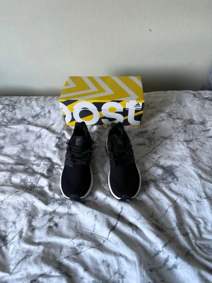 Svarta Adidas Ultra Boost sneakers - Säljer ett par svarta Adidas Ultra Boost sneakers med vit sula och klassisk Boost-dämpning. Ovandelen är i mesh för bra andningsförmåga och skorna har svarta snören samt en stabil hälkappa. Perfekta för dig som gillar stilrena och bekväma sneakers.