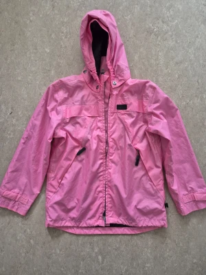 Vindjacka - Snygg rosa vindjacka med huva och dragkedja framtill. Jackan har två snedställda fickor med dragkedja och justerbara muddar vid ärmsluten. Materialet är tunt och vindtätt, perfekt för blåsiga dagar. Enkel och stilren design utan synligt märke. Några fläckar.