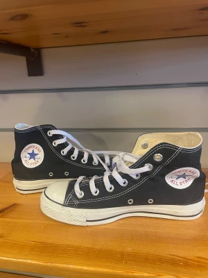 Svarta Converse All Star Highs - Klassiska svarta Converse All Star sneakers med högt skaft, vita gummisulor och tåhätta. Skorna har vita snören och tydliga logopatchar på sidan. Tillverkade i canvas med kontrastsömmar och metallöljetter.