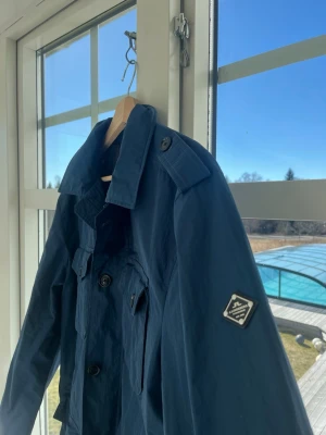 Mörkblå field jacka från J.Lindeberg - Snygg mörkblå vindjacka från J.Lindeberg med klassisk krage, axelklaffar och flera fickor framtill. Jackan har en patch med logga på ärmen och stängs med både dragkedja och knappar. Perfekt för blåsiga dagar och stilren look. Även perfekt nu till lite varmare vår väder! 