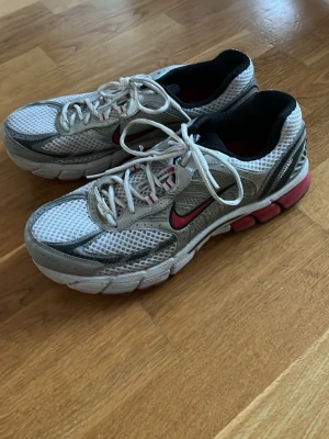 Nike vita och grå sneakers med röd swoosh - Nike sneakers i vitt och grått med mesh-ovandel och röda detaljer, inklusive den klassiska swoosh-loggan. Skorna har vita snören, svart vadderad insida och en robust sula med röda inslag. Perfekta för dig som gillar sportig och retro stil.