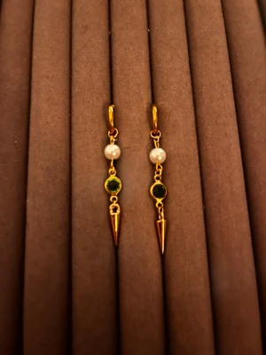 Guldörhängen med pärlor och grön sten - Säljer ett par unika guldtonade örhängen med vita pärlor, grön rund sten och spetsiga hängen. Örhängena har en cool och modern design med mix av material som ger en edgy känsla. Perfekta för dig som vill sticka ut med dina accessoarer.