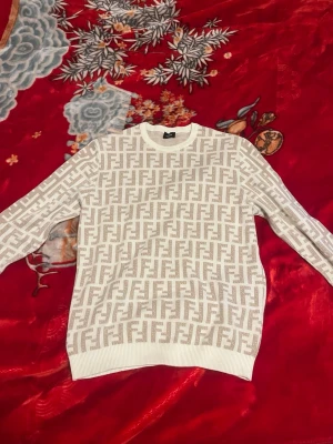 Vit stickad tröja från Fendi - Snygg vit stickad tröja från Fendi med det klassiska FF-monogrammet i beige över hela plagget. Tröjan har rund halsringning, långa ärmar och ribbade muddar vid ärmslut och nederkant. Perfekt för dig som gillar exklusiv och trendig design.