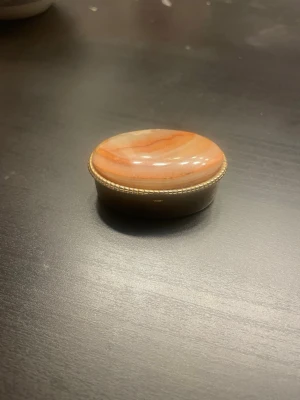 Oval liten smyckesask i metall och sten - Säljer en liten oval smyckesask med lock i polerad sten i orangea och vita toner. Asken har en dekorerad metallkant och graverad metall runt om. Perfekt för småsaker eller smycken.