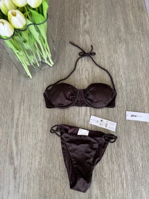 Mörkbrun bikini från Gina Tricot - Snygg mörkbrun bikini från Gina Tricot med bandeau-topp och knyt i nacken. Underdelen har smala band i sidorna och båda delarna har en stilren, enkel design. Perfekt för lata dagar på stranden eller poolen. Helt ny och oanvänd, köpt för 440kr 