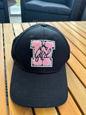 Svart keps med rosa patch / Y2k - Snygg svart keps med böjd skärm och en stor rosa patch med bokstaven 'W' och svart broderad text på framsidan. Kepsen har klassisk form och är tillverkad i bomullstyg. Perfekt accessoar för en trendig och avslappnad stil.