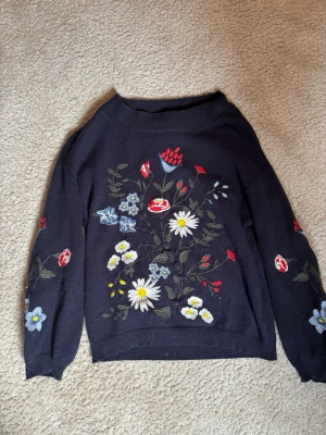 Mörkblå stickad tröja med blommor - Mysig mörkblå stickad tröja med broderade blommor i vitt, rött, blått och gult på både fram- och ärmar. Tröjan har rund halsringning och ribbade muddar. Perfekt för dig som gillar färgglada detaljer. Strl 134 men skulle säga att den passar jättebra på mig som är strl XS/S. Passar sååå bra på sommaren och är otroligt bekväm🩵