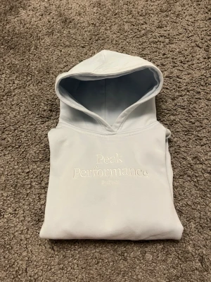 Ljusblå hoodie Peak Performance - Snygg ljusblå hoodie från Peak Performance med broderad logga på bröstet. Tillverkad i mjukt material med borstad insida för extra komfort. Klassisk huva och stilren design, perfekt för en avslappnad look.