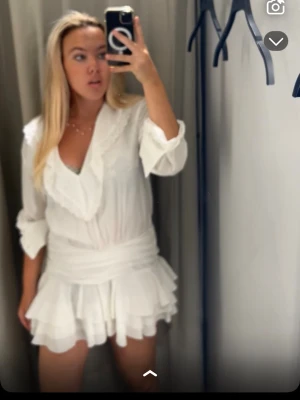 Vit volangklänning med v-ringning - Supersöt vit klänning med v-ringning och volangdetaljer både vid halsen och längs den korta kjolen. Klänningen har långa ärmar och ett omlottliknande liv. Perfekt för sommarens alla tillfällen!