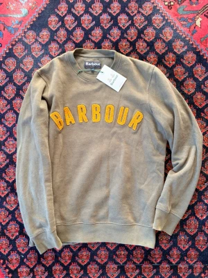 Olivgrön sweatshirt från Barbour - Säljer en olivgrön sweatshirt från Barbour med stora gula bokstäver i frotté på bröstet. Tröjan har rund halsringning, ribbade muddar och är tillverkad i mjuk bomullsblandning. Perfekt för en avslappnad och trendig stil.