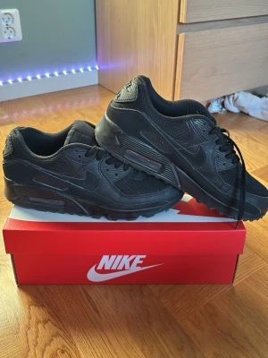 Nike Air Max 90 helsvarta sneakers - Säljer ett par helsvarta Nike Air Max 90 sneakers med klassisk chunky sula och diskret Nike-logga på sidan och hälen. Skorna är tillverkade i en mix av mesh och syntetmaterial för en sportig look. Perfekta för dig som gillar stilrena och tidlösa sneakers.