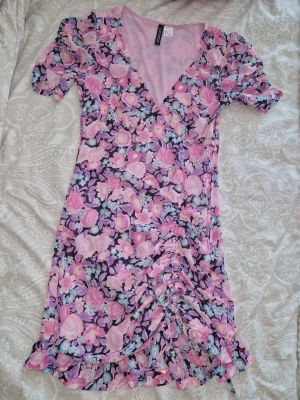 Blommig kortklänning från HM  Divided - Cute kortklänning från HM med blommigt mönster i rosa, lila och blå toner. Klänningen har v-ringning, korta puffärmar och rynkade detaljer framtill. Perfekt för sommarens alla tillfällen!