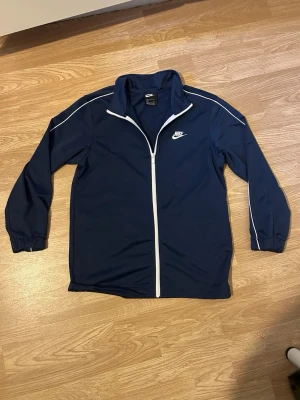Marinblå Nike tröja - Snygg marinblå träningsjacka från Nike med vit dragkedja och vita detaljer längs axlar och ärmar. Jackan har hög krage, långa ärmar och klassisk Nike-logga på bröstet. Perfekt för träning eller sportiga outfits.