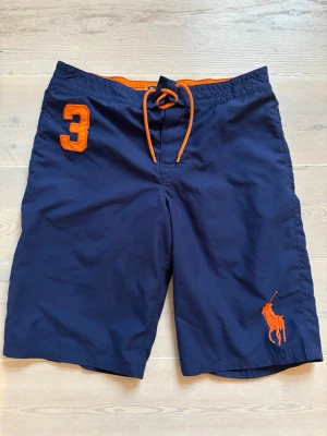 Marinblå badbyxor från Polo Ralph Lauren - Snygga marinblå badbyxor från Polo Ralph Lauren med orange detaljer, inklusive snörning, stort broderat nummer 3 och klassiska polospelaren-loggan. Perfekta för stranden eller poolen, tillverkade i snabbtorkande material. Storlek 18-20 år