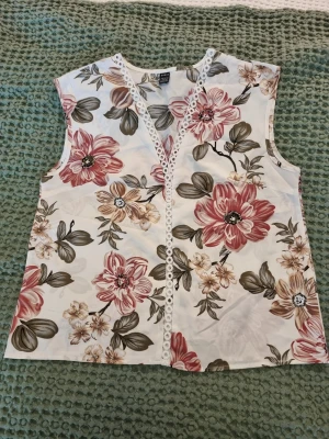 Blommig ärmlös topp från SHEIN - Snygg ärmlös topp från SHEIN med stort blommigt mönster i rosa, beige och grönt på vit botten. V-ringad med dekorativ spets längs urringningen. Perfekt för dig som gillar romantisk och somrig stil.