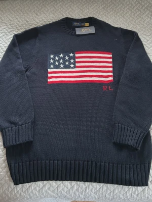 Mörkblå stickad tröja Polo Ralph Lauren - Snygg mörkblå stickad tröja från Polo Ralph Lauren med amerikanska flaggan broderad på bröstet och RL-broderi i rött. Tröjan har rund hals, ribbade muddar och nederkant. Perfekt för dig som vill ha en klassisk och stilren look.
