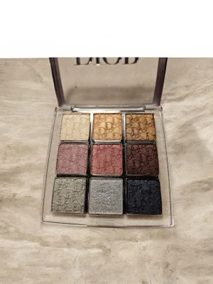 Dior ögonskuggspalett 9 nyanser - Snygg ögonskuggspalett från Dior med 9 olika nyanser i metallic och skimrande finish. Färgerna går i guld, brons, vinröd, lila, silver, grått och mörkblått. Paletten har fyrkantiga skuggor med Dior-logga präglad i varje ruta och genomskinligt lock.