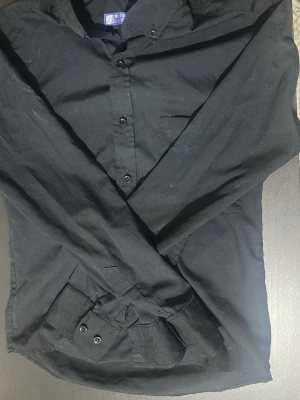 Svart skjorta från Boudkian - Klassisk svart skjorta från Boudkian med lång ärm och knappar framtill. Skjortan har en enkel bröstficka och stilren design, perfekt för dig som gillar en clean och tidlös look. Tillverkad i mjukt material som känns bekvämt hela dagen.