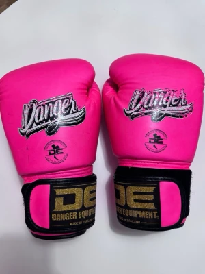 Boxnings tillbehör  - Snygga rosa boxningshandskar från Danger Equipment med svart och guld logga på handledsremmen. Tillverkade i semi-läder och har kardborreknäppning för enkel justering. Perfekta för träning inom boxning eller kampsport. Även vita benskydd från unbreakable i bra skick. (Grenskydd oanvänt ingår)