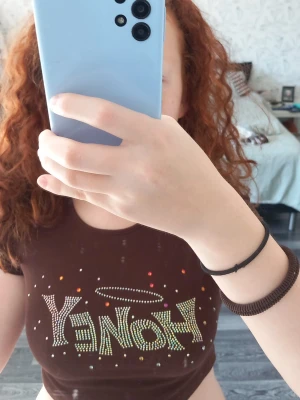 Brun croppad topp med texten HONEY - Snygg brun croppad topp med korta ärmar och texten 'HONEY' i glittriga stenar framtill. Toppen har färgglada detaljer och ett litet hål i ryggen vid nacken. Perfekt för dig som vill sticka ut med en trendig och lekfull stil.