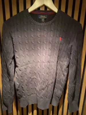 Mörkblå kabelstickad tröja Polo - Kabelstickad mörkblå tröja från Polo Ralph Lauren med rund halsringning och röd broderad logga på bröstet. Storleken är L (14-16) men motsvarar storlek S i vuxen storlek Tröjan har ribbade muddar vid ärmslut och nederkant, och är tillverkad i mjukt material som passar perfekt till höst och vinter.
