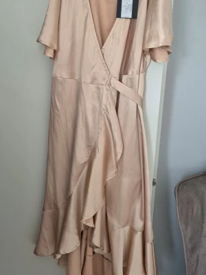 Champagnefärgad omlottklänning - Elegant långklänning i champagnefärgad satin med omlottmodell och volangdetaljer. Klänningen har korta ärmar och ett knytband i midjan för en feminin siluett. Perfekt för dig som vill ha en stilren och lyxig look.