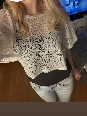 Vit spetsblus med blommönster - Säljer en vit, croppad spetsblus med blommigt mönster och vida, korta ärmar. Blusen har en luftig känsla och är perfekt att styla över ett linne eller topp. Supersnygg till jeans eller kjol för en trendig look.(köp med Instabox)