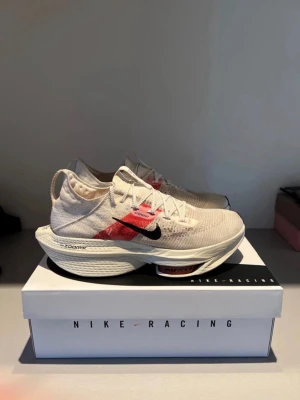 Nike ZoomX Vaporfly Next% 2 springskor  - Nike ZoomX Vaporfly Next% 2 sneakers i beige med röda och svarta detaljer. Ovandel i mesh för bra ventilation, svart swoosh-logga på sidan och ZoomX-mellansula för extra studs. Lätta och sportiga med modern design, perfekta för dig som gillar att springa snabbt.