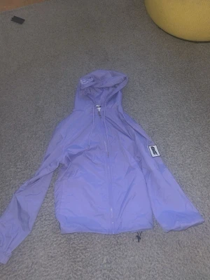 Lila vindjacka från Risky - Snygg lila vindjacka från Risky med huva och dragkedja framtill. Jackan har en patch på ärmen och justerbar dragsko nedtill. Perfekt för blåsiga dagar och streetwear-stil.