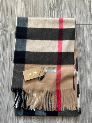 Burberry klassisk rutig halsduk - Säljer en ikonisk halsduk från Burberry i klassiskt rutigt mönster med beige, svart, vitt och röda detaljer. Halsduken är tillverkad i 100% kashmir och har fransar i ändarna. Perfekt accessoar för att lyfta din outfit med en tidlös och lyxig känsla.