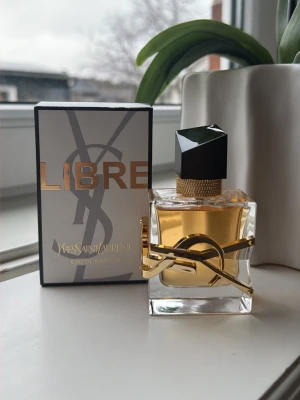 Yves Saint Laurent Libre EdP - Yves Saint Laurent Libre Eau de Parfum i en elegant, fyrkantig glasflaska med gulddetaljer och svart asymmetrisk kork. Doften kommer i originalkartong med guldtext och YSL-logga. Parfymen har en lyxig och modern design som sticker ut på hyllan.
