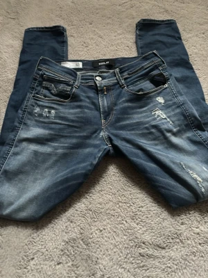 Blå slitna jeans från Replay - Säljer ett par blå jeans från Replay med slitningar och tvättade detaljer. Klassisk femficksmodell med knappgylf och läderpatch bak i midjan. Jeansen har en modern passform och är tillverkade i denim med snyggt sliten look. De har en storlek på 28x32, priset kan diskuteras vid snabbaffär