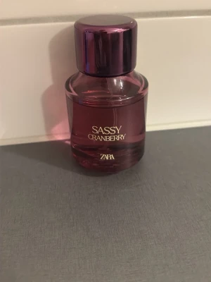 Zara Sassy Cranberry parfym - En snygg parfymflaska från Zara med doften Sassy Cranberry. Flaskan är rund och har en djup bärig rosa färg med matchande lock. Texten på flaskan är i guld och designen känns modern och ungdomlig. Perfekt för dig som gillar fruktiga dofter.