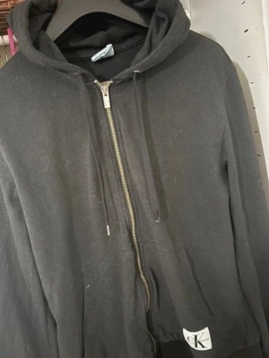 Svart hoodie från Calvin Klein - Snygg svart hoodie från Calvin Klein med dragkedja framtill och klassisk CK-logga på ärmen. Hoodien har huva med dragsko och känguruficka. Tillverkad i mjukt bomullsmaterial som är skönt att bära.