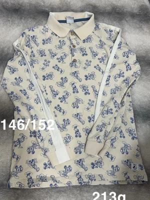 Långärmad topp med Musse Pigg-mönster - Långärmad topp från polarn o pyret i ljusbeige med blått Musse Pigg-mönster över hela plagget. Klassisk krage, knappar framtill och ribbade muddar vid ärmslut. Perfekt för dig som gillar Disney och vill ha en lekfull och unik stil.🌸 Fläck på ena mudden. Storlek 146/152. 