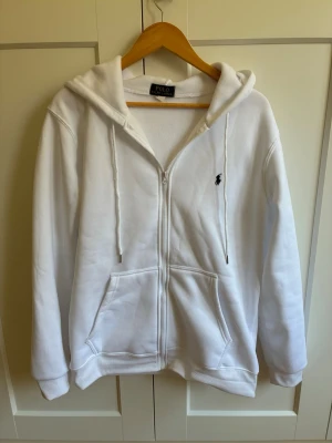 Vit hoodie från Polo Ralph Lauren - Klassisk vit hoodie med dragkedja från Polo Ralph Lauren. Har huva med dragsko, två stora fickor framtill och den ikoniska lilla svarta loggan broderad på bröstet. Extremt skönt material.