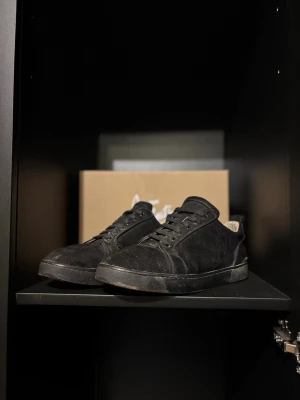 Svarta sneakers från Christian Louboutin - Stilrena svarta sneakers i mocka från Christian Louboutin med klassisk röd sula och lågt skaft. Skorna har svarta snören och diskreta sömmar som ger en clean look. Perfekta för dig som vill ha något exklusivt och tidlöst.