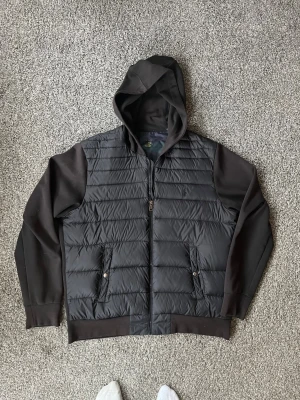 Svart cardigan med huva från Polo Ralph Lauren - Snygg svart cardigan från Polo Ralph Lauren med huva och dragkedja framtill. Jackan har quiltad front, mjuka ärmar och två fickor med knappstängning. Perfekt för kalla vinterdagar och riktigt stilren look.