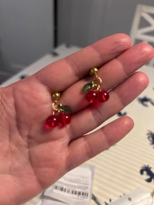 Stål Guldörhängen med körsbärspärlor - Säljer ett par örhängen med stål  guldtonad metall och röda körsbärspärlor i glas  Örhängena har små gröna blad i som detalj och bärformade hängen som ger en söt och lekfull look.