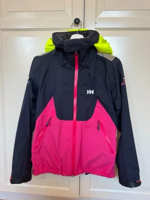 Helly Hansen vindjacka svart/rosa - Snygg vindjacka från Helly Hansen i svart och starkt rosa med detaljer i neon-gult på huvan. Jackan har dragkedja framtill, två fickor med dragkedja och justerbar huva. Perfekt för blåsiga  dagar och outdoor-aktiviteter. I fint skick! Skriv för frågor eller fler bilder💕💕Nypris är runt 3000 kr, priset kan diskuteras!